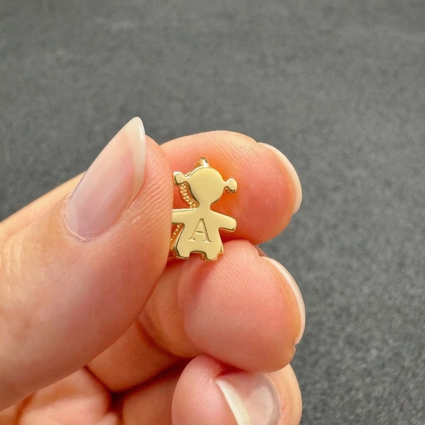 Gold girl-shaped pendant with a letter 'A' held between fingers against a gray background