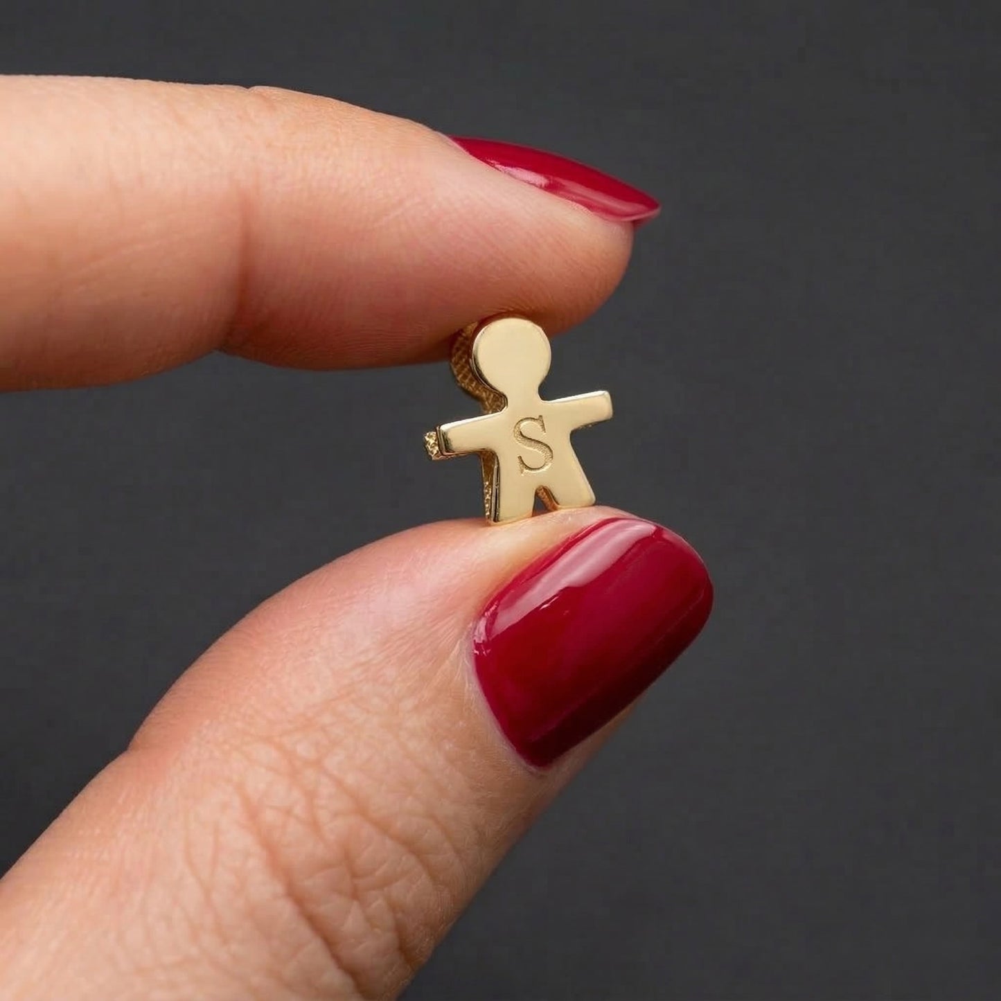 Gold charm shaped like a person with initials held between fingers against a dark background