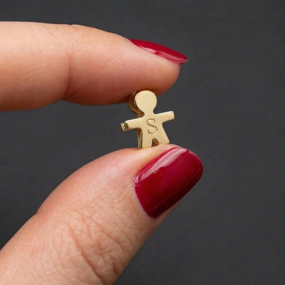 Gold charm shaped like a person with initials held between fingers against a dark background