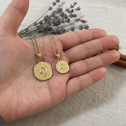 Two gold necklaces with pendants held in a hand against a neutral background.